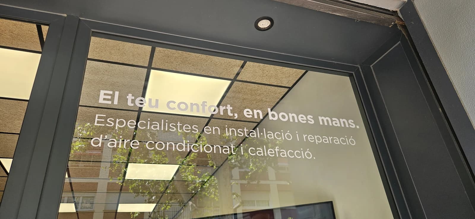 Texto blanco de vinilo sobre el cristal de un negocio de climatización, con reflejos del exterior.
